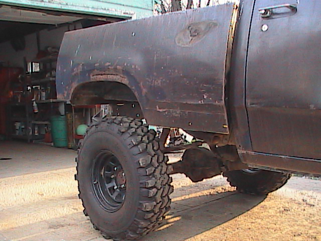 Dark-colored truck bed and large off-road tire, showing rust and damage. Visible drivetrain components and a garage interior