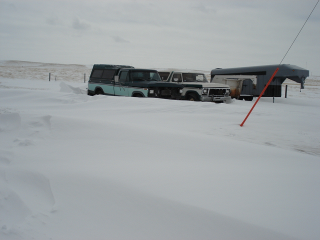 Two trucks and a trailer partially buried in snowdrifts under a cloudy sky