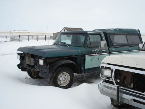 1979 Ford F-150 - Two older model pickup trucks are parked in a snowy field with farm buildings in the background.