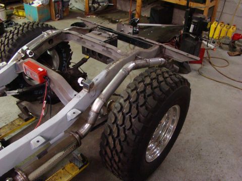 1948 Jeep Wrangler - The chassis of a Jeep is shown with large off-road tires, a custom exhaust system, and a red ignition control module.
