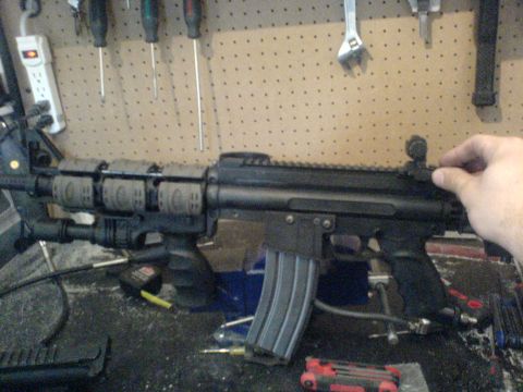 1985 Ford Bronco - A person is holding the rear sight of an assault rifle on a workbench with tools hanging on a pegboard.
