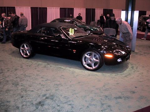 1996 Ford Taurus - A black convertible sports car is displayed indoors at an auto show with other vehicles and people present.