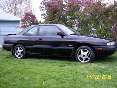1989 Mazda MX-6 - A black 1989 Mazda MX-6 GT coupe is shown from the side, parked on a grassy lawn with a house and trees in the background.