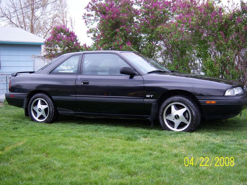 Black 1989 Mazda MX-6 GT coupe parked on grass, side view. Aftermarket wheels and a spoiler are visible