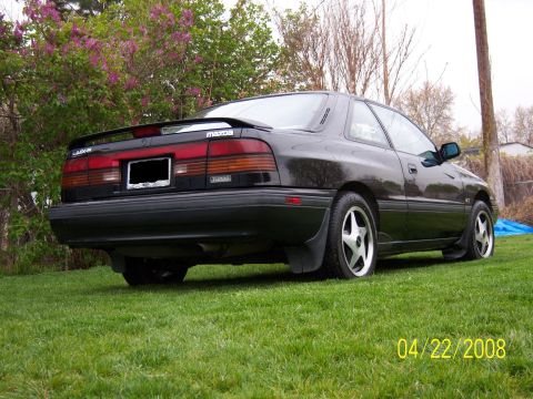 1989 Mazda MX-6 - A 1989 Mazda MX-6 is shown from the rear quarter angle, parked on a grassy lawn.