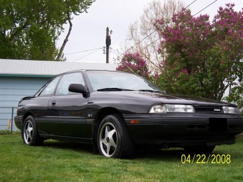 1989 Mazda MX-6 - A black 1989 Mazda MX-6 GT coupe is shown from the front 3/4 angle on a grassy lawn.