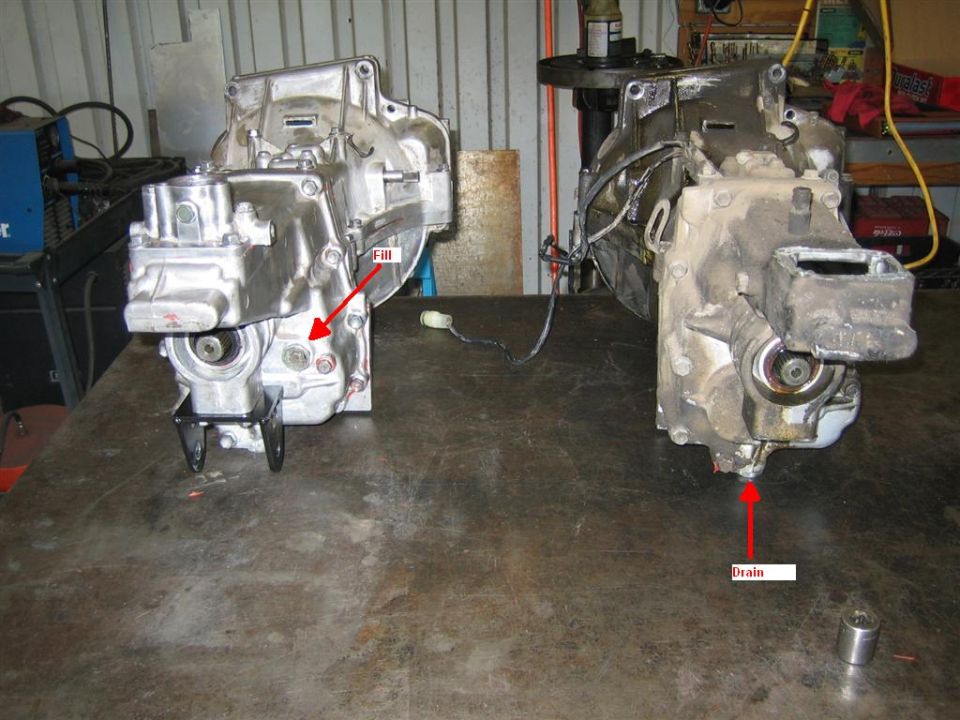Two aluminum transmissions on a metal table, one labeled 'Fill' and the other with a 'Drain' label pointing to its underside
