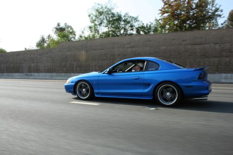 1992 Ford Crown Victoria - A blue coupe, likely a 1994 Ford Mustang, cruises down the highway on a clear day.