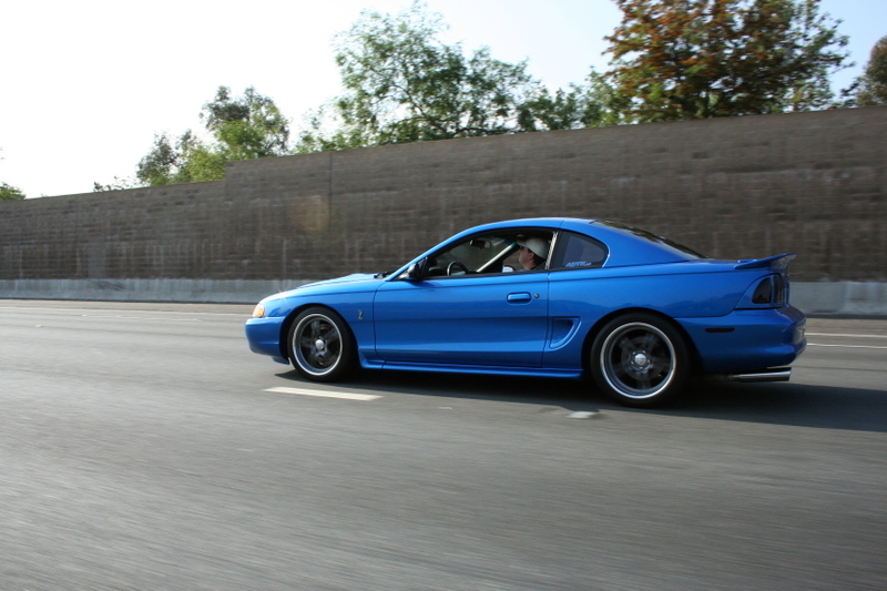 Blue coupe driving on highway, side view. Appears to be a 1994 Ford Mustang