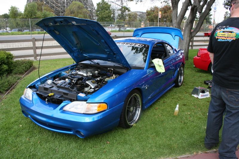 Blue Ford Mustang with hood and trunk open, parked on grass. A red car is visible in the background