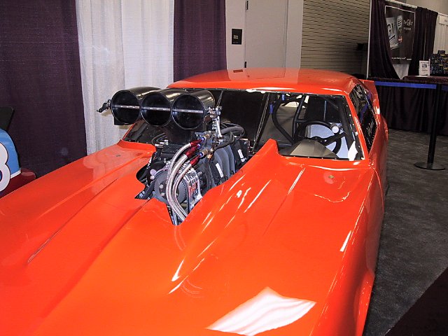 Orange drag car with a large blower protruding from the hood, showing engine components and roll cage