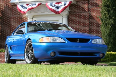 1992 Ford Crown Victoria - A blue 1996 Ford Mustang Cobra is parked on a grassy lawn in front of a brick building with patriotic bunting.