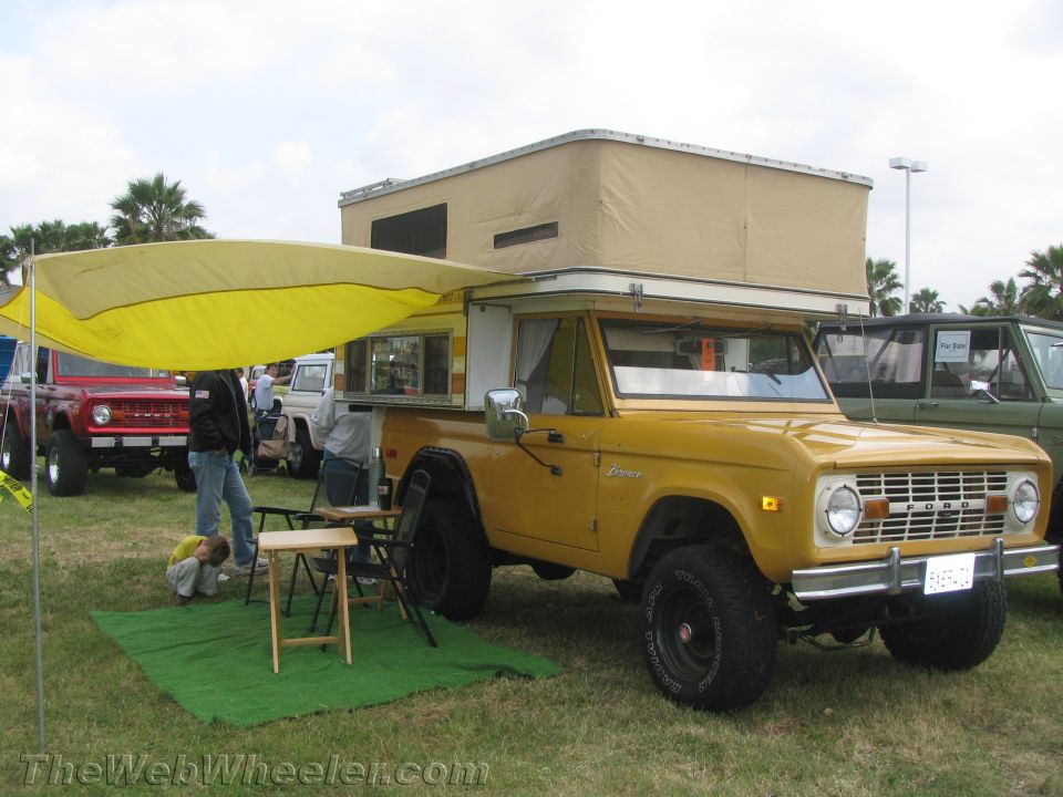Yellow Ford Bronco with camper shell and awning set up, parked next to a red Bronco
