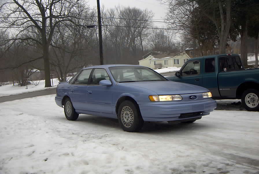 Light blue Ford Taurus sedan with headlights on, parked in snow, front 3/4 view