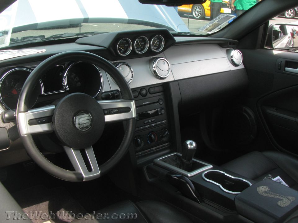 Interior of a Ford Mustang showing the dashboard, steering wheel, center console, and gear shifter. A Cobra emblem is visible on the center console lid