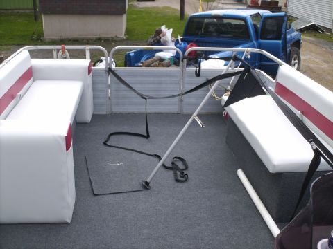 Pontoon boat interior with white seats and gray carpet, a blue pickup truck with open doors is parked behind…