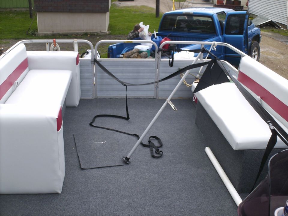 Pontoon boat interior with white seats and gray carpet, a blue pickup truck with open doors is parked behind it