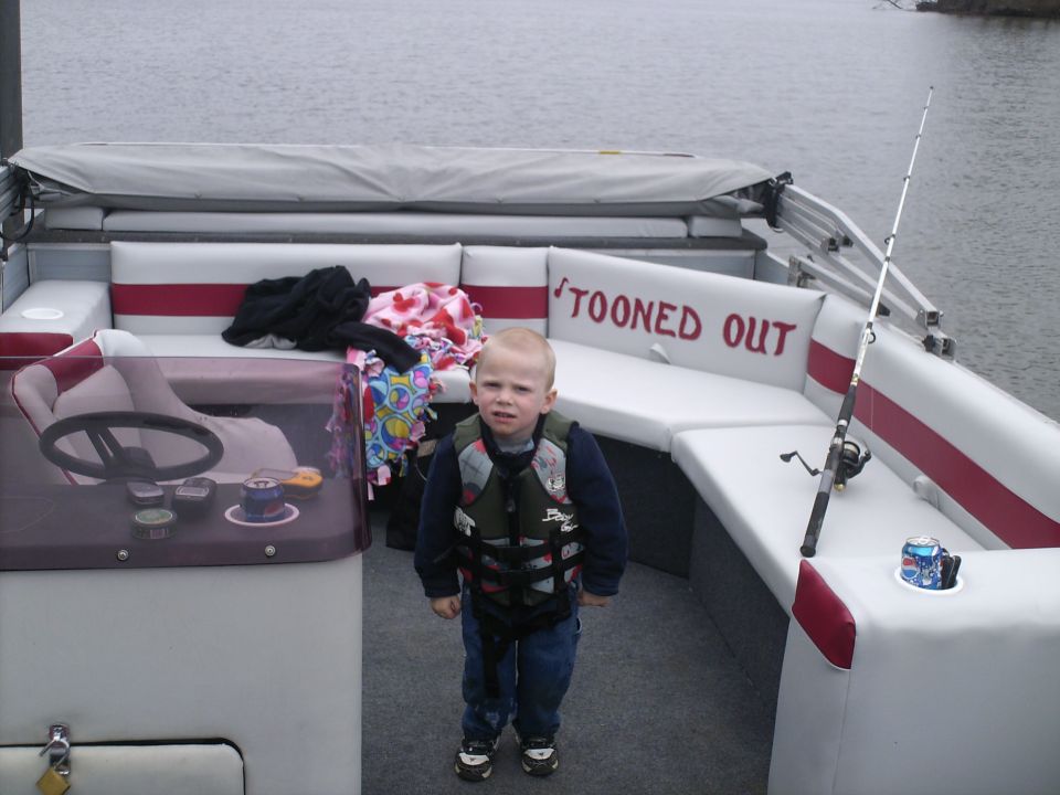 Child wearing a life vest stands on a boat with white seats and red trim. A fishing rod is visible