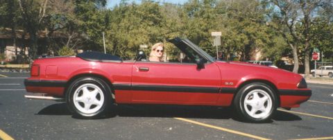 1991 Ford Mustang - A red 1991 Ford Mustang convertible is seen from the side, with the top down and a woman driving.