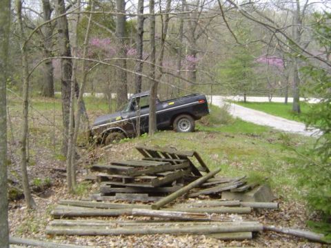 1979 Ford Bronco - Wide shot of a black Ford truck nosed down off the edge of a gravel drive into a wooded ditch, rear wheel raised off the ground. Stacked wo…