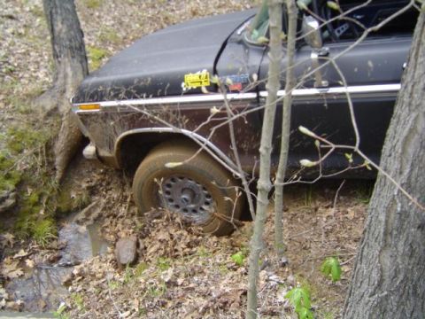 1979 Ford Bronco - Passenger-side view of a black and brown two-tone Ford truck stuck at an angle in a muddy wooded bank, front tire wedged into the dirt. Chr…