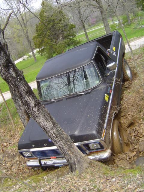 1979 Ford Bronco - Black Ford truck tipped nose-down into a muddy ditch beside a gravel drive, front passenger wheel buried and body leaning against a tree. F…