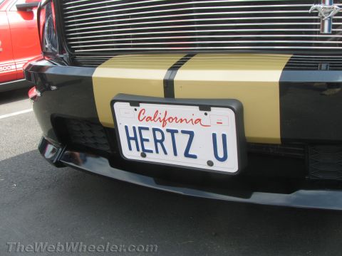 1968 Ford Mustang - A black Ford Mustang with gold racing stripes is parked next to a red vehicle. The Mustang's front bumper and license plate are visible.
