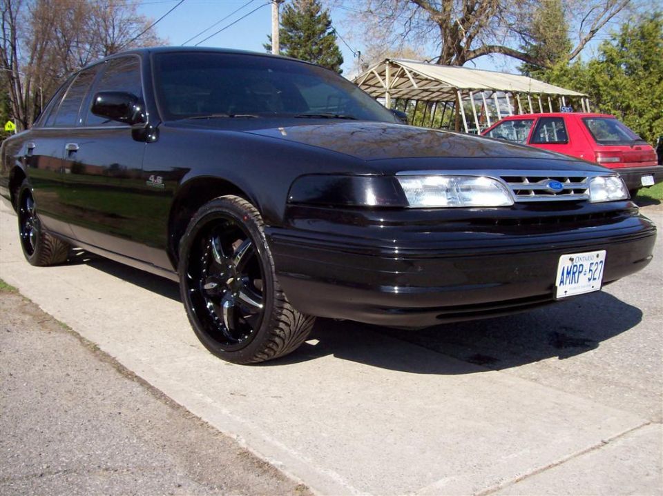 Black Ford Crown Victoria with chrome rims parked on a concrete driveway. Red car visible in background