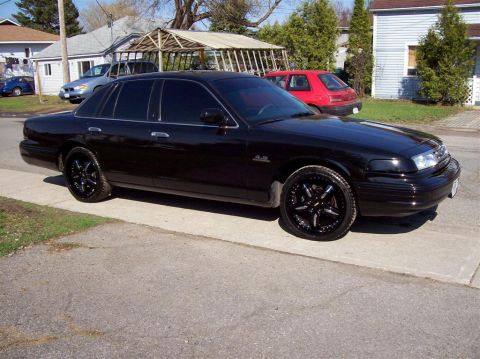 1996 Ford Crown Victoria - A black sedan with aftermarket rims is parked on a driveway, with a red hatchback in the background.