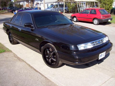 1996 Ford Crown Victoria - A black sedan with custom wheels is parked on the street, with another red car visible in the background.