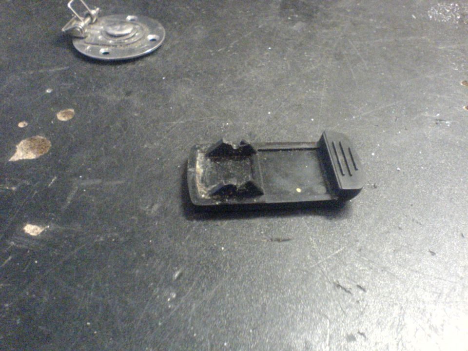Black plastic car part with clips on a dark surface. A metal circular piece with a latch is nearby