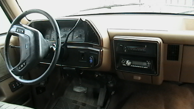 Tan dashboard and steering wheel of a 1987 Ford F-150 truck, showing aftermarket radio and floor mats