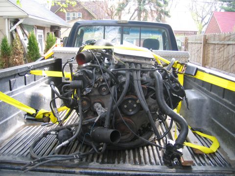 1996 Ford Crown Victoria - Engine being transported in the bed of a pickup truck, secured with yellow tie-down straps.