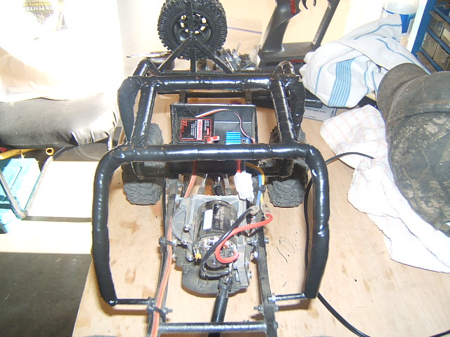 RC crawler chassis with exposed motor, battery pack, and roll cage. Spare tire mounted on top