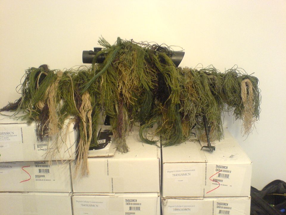 Sniper ghillie suit draped over boxes, with a scope visible on top