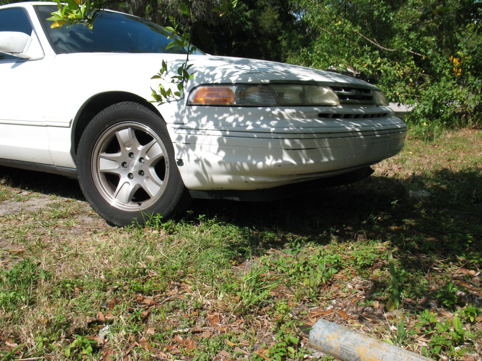 White sedan, front view, parked outdoors with foliage overhead. Visible damage on front bumper