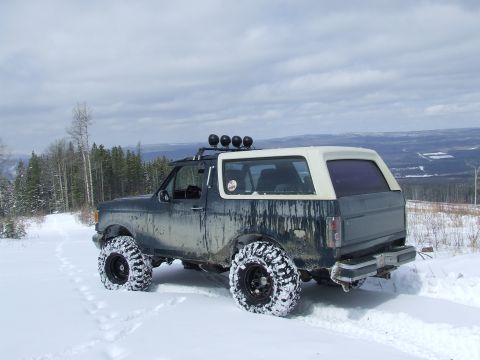 1988 Ford Bronco - Rear three-quarter view of a lifted dark green Ford Bronco with a cream fiberglass top, parked in deep snow on a ridge above a snow-covered…