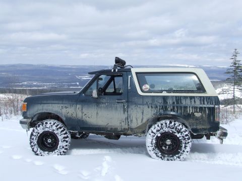 1988 Ford Bronco - Driver-side profile of a lifted dark green full-size Ford Bronco with a weathered white fiberglass top, parked in deep snow on a ridge over…