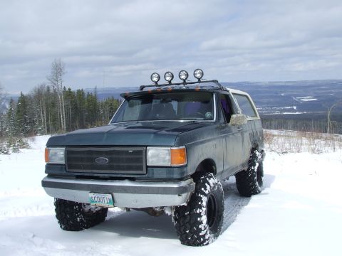 1988 Ford Bronco - Front three-quarter view of a lifted dark green full-size Ford Bronco with a cream fiberglass top, parked in snow on an open ridge with spr…
