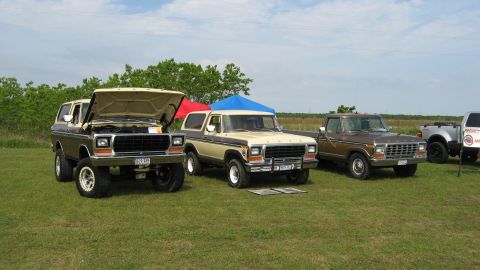 1979 Ford Bronco - Multiple Ford Broncos and a pickup truck are parked on a grassy field under a partly cloudy sky.