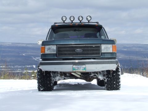 1988 Ford Bronco - Head-on view of a lifted dark green Ford Bronco standing on a snow-covered ridge, four round driving lights on a roof light bar and a whip…