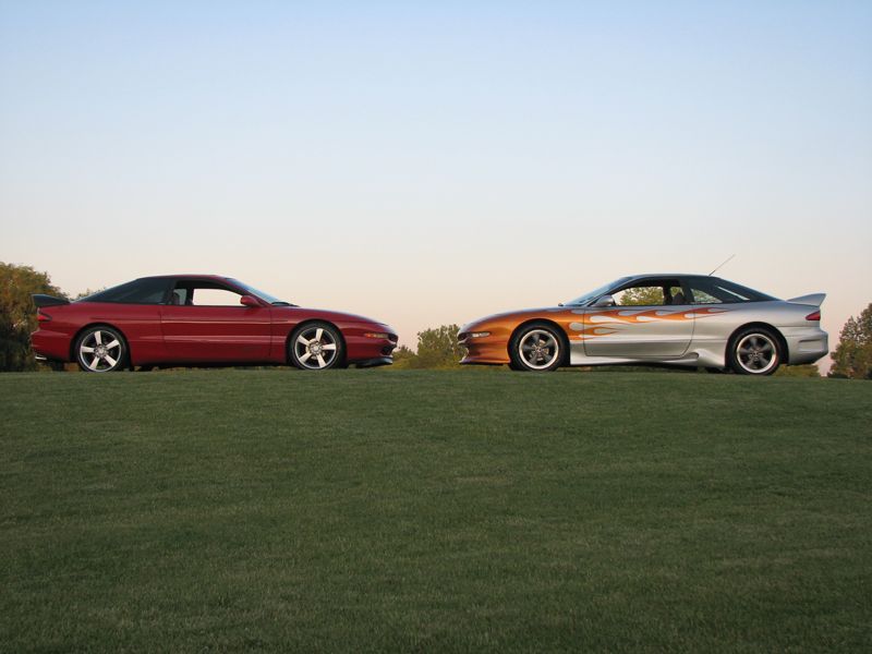 Two 1993 Ford Probes parked on a grassy hill, one red, one silver with flames