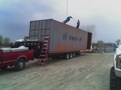 1992 Ford Bronco - A red pickup truck is parked next to a large shipping container that is being loaded by workers.