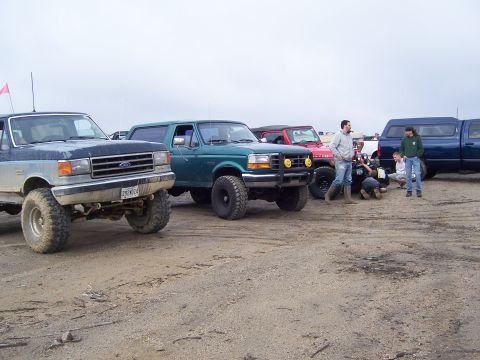 1996 Ford Bronco - Several off-road vehicles, including a 1996 Ford Bronco, are parked on a dirt lot with people gathered.