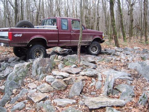 1996 Ford Bronco - A maroon Ford F-Series pickup truck navigates a challenging rocky terrain in a wooded area.