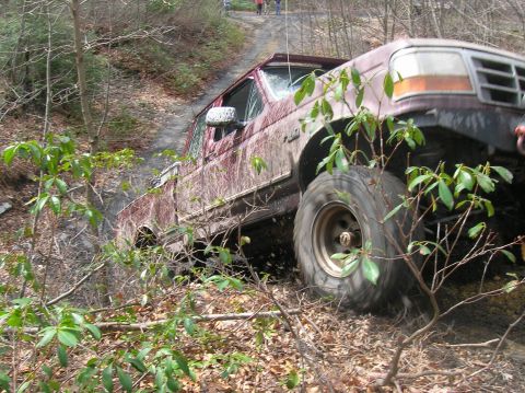 1996 Ford Bronco - A maroon Ford F-Series truck navigates a muddy, off-road trail surrounded by trees and foliage.