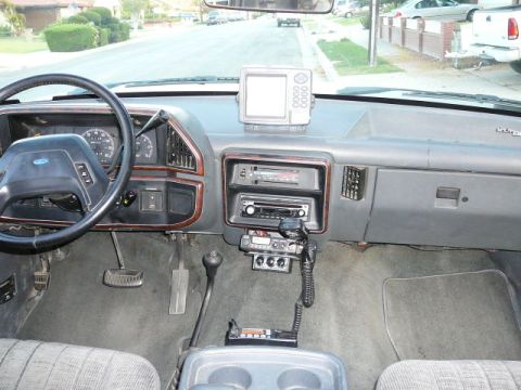 1988 Ford Bronco - Interior view from the rear seat of a gray Ford truck dashboard with a wood-grain-trimmed center stack holding a stacked head unit and cass…