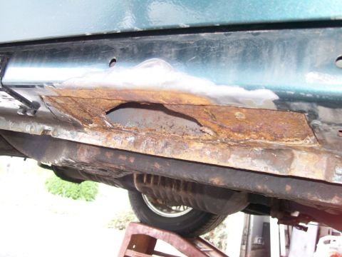 1995 Mercury Grand Marquis - Low-angle shot under a green car raised on a jack stand, showing the rocker panel and floor pan area cut open to expose heavy rust with a j…