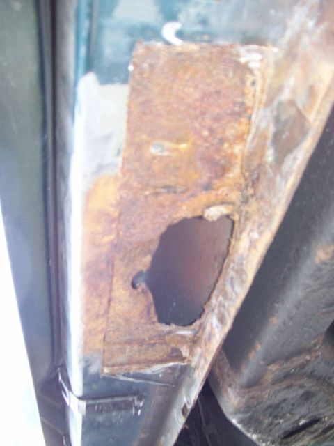 1995 Mercury Grand Marquis - Close-up of severe rust damage at a lower door or rocker area on a dark green car, with the paint stripped back to bare metal and a jagged…