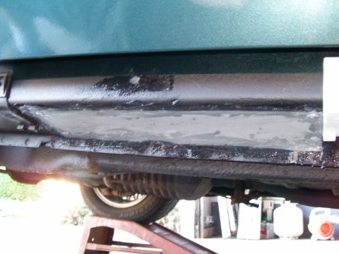 1995 Mercury Grand Marquis - Underside view of a green car on a jack stand showing a rust-repair patch panel welded into the rocker/floor area and skimmed with gray bod…